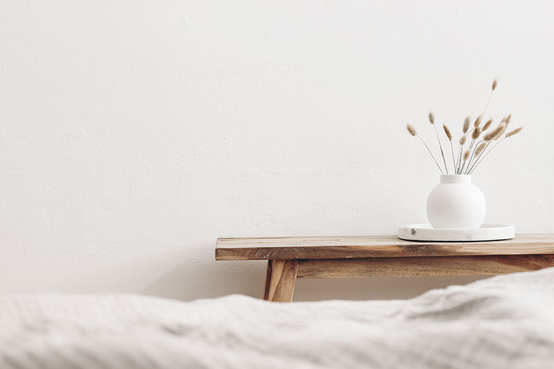 A minimalist scene with a wooden bench against a white wall, holding a round white vase filled with dried grass on a white tray. The foreground shows the edge of a soft, light-colored blanket.