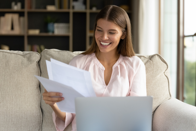 A smiling woman sits on a couch holding papers in one hand while looking at a laptop, appearing happy and relaxed in a cozy, well-lit living room with bookshelves in the background.