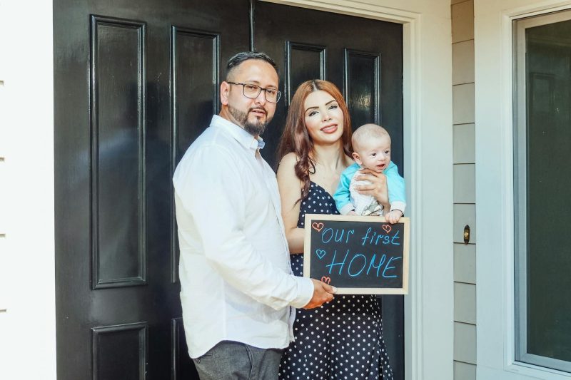 Home 13 Young family with baby holding a sign Our First Home at door 1774371664